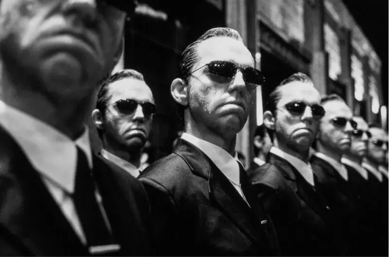 Fila di uomini identici in abito scuro e occhiali da sole, simbolo del conformismo nella narrativa dominante sull'AI